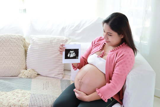 Happy smiling young asian pregnant woman resting and sitting on sofa in living room while showing ultrasound image. Expectant mother preparing and waiting for baby birth during pregnancy.