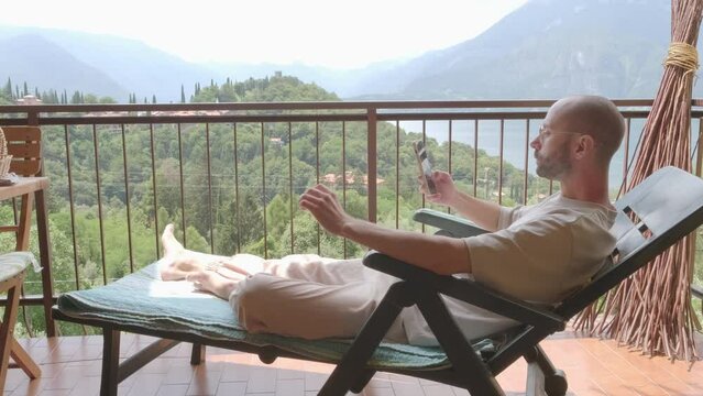 positive charming young man using modern phone, lying relaxed on lounger on balcony against backdrop of mountains, highlighting role of gadgets in everyday business communication, Digital Lifestyle