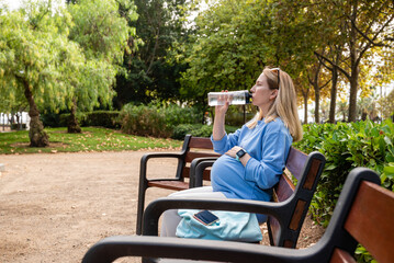 Relaxed athletic pregnant woman, sits on a park bench and drinks water. Outdoor fitness training