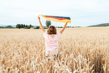 A happy girl stands in a field of spikelets at sunset with the German flag.