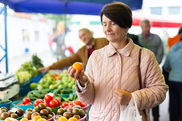 Smiling elderly female customer choosing organic oranges while walking at local bazaar