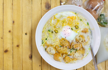 Hot congee or Rice porridge with soft boiled egg served in a white bowl with deep-fried dough stick (crispy patongo) on woodden backgrounds,left copy space fortext.