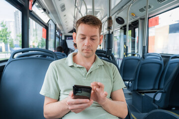 Attractive man is reading emails on his smartphone connected to public wi-fi while sitting in a city bus. Handsome male is looking at the screen of a mobile phone while texting message