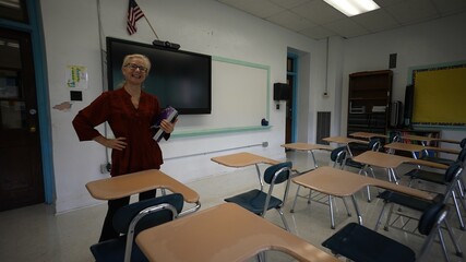 Portrait of smiling happy teacher entering an empty classroom, turning lights, on optimistic about the future of education. US American flag and smart board in room.