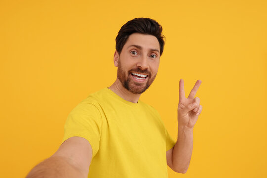 Smiling man taking selfie and showing peace sign on yellow background