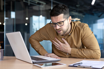 Heart attack, panic attack at work. A young man holds his chest while sitting at the desk in the office. Winced from pain, feels discomfort