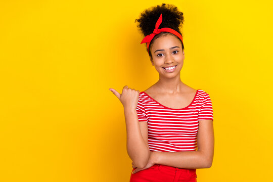 Photo of adorable cheerful girl dressed red striped t-shirt pointing thumb empty space isolated yellow color background