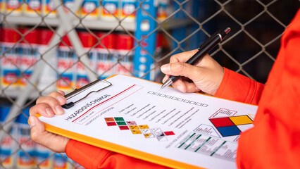 A worker is checking on the hazardous chemical material information form with background of chemical storage area at the factory place. Industrial safety working action. Selective focus. - obrazy, fototapety, plakaty