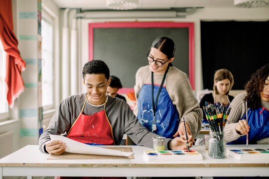 Smiling male teenage student showing artwork to female teacher during painting class