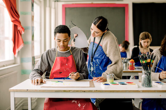 Young Female Teacher Guiding Male Teenage Student During Art Class At High Class