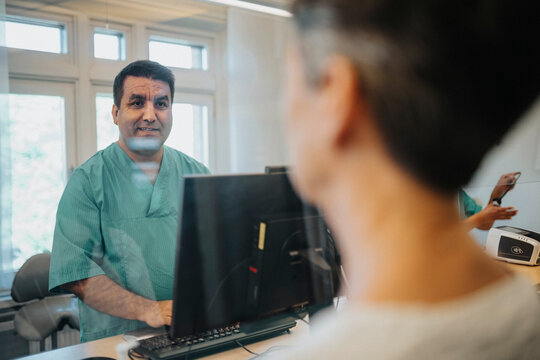 Male receptionist talking with patient through glass screen at reception desk in hospital