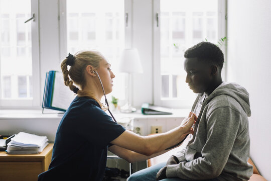 Side view of female healthcare worker checking heartbeat of teenage male patient sitting in clinic