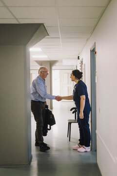 Female nurse doing handshake with male senior patient while standing in corridor at hospital