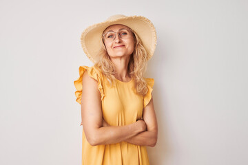 Portrait of happy beautiful blonde mature woman in yellow summer dress, glasses and straw hat crossed arms smiling joyfully isolated on white studio background. Vacation and travel concept