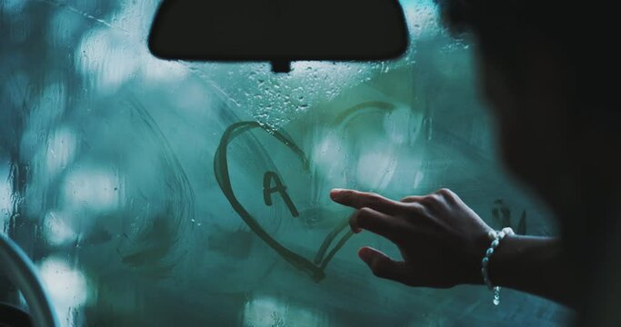 Man drawing a heart shape with initials on a car window.
