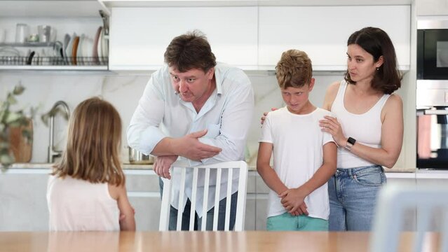 At home in kitchen, parents scold their daughter for careless act. Sulky preschool girl stands with arms crossed in front of her, parents during serious conversation. Mom hugs son