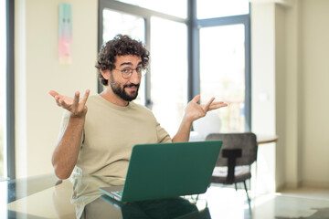 young adult bearded man with a laptop feeling puzzled and confused, doubting, weighting or choosing...