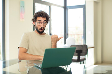 young adult bearded man with a laptop looking astonished in disbelief, pointing at object on the side and saying wow, unbelievable