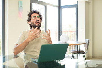 young adult bearded man with a laptop looking desperate and frustrated, stressed, unhappy and annoyed, shouting and screaming