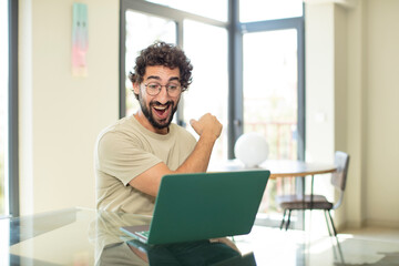young adult bearded man with a laptop feeling happy, positive and successful, motivated when facing a challenge or celebrating good results