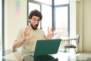 young adult bearded man with a laptop feeling stupefied and scared, fearing something frightening,...