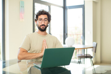 young adult bearded man with a laptop feeling worried, hopeful and religious, praying faithfully...