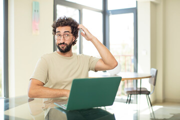 young adult bearded man with a laptop feeling puzzled and confused, scratching head and looking to the side