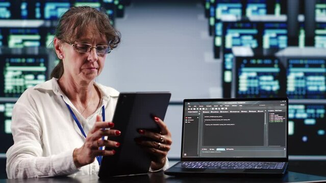 Elderly woman working in IT in high tech workplace completing tasks on tablet while running scripts on laptop terminal. Professional amidst server cabinets manipulates code and improves cybersecurity