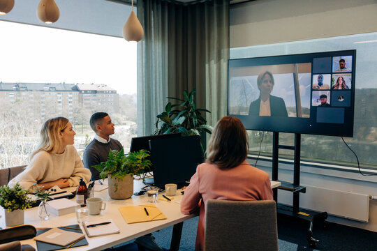 Male and female colleagues doing video call with businesswoman over TV in coworking office