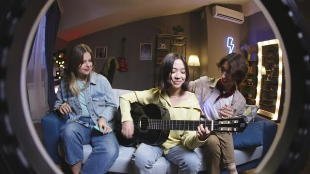 Three friends filming through ring lamp, they play guitar and have fun sitting on couch. Life in social media and recreation applications, content creators in funny cringe mood recording video content