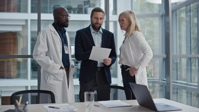 Medium shot of doctors and manager of clinic standing in room with panoramic windows, looking at document and discussing results of year
