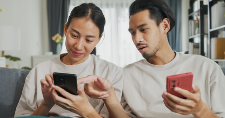 Young Asian couple sit on couch spend time together have fun use smartphone devices online shopping at home. Happy Young husband and wife laugh relax with phone, Lifestyle domestic activity concept.