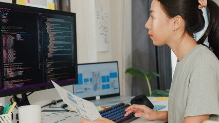 Side view of professional young Asia girl IT development programmer typing on keyboard coding programming fixing data code on computer screen and laptop on table in workroom at house office.