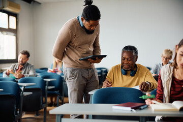 Black adult education teacher assists senior student with assignment during class in classroom.