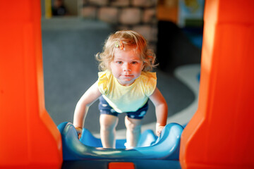 Happy blond little toddler girl having fun and sliding on indoor playground at daycare or nursery....