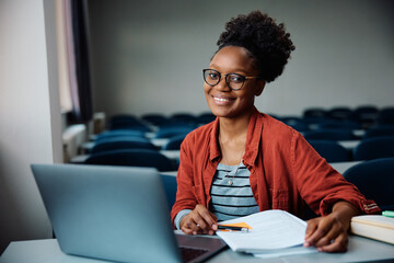 Happy black female student using laptop while learning in classroom and looking at camera.