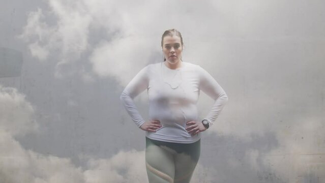 Animation of clouds over plus size caucasian woman in sports clothes flexing muscles