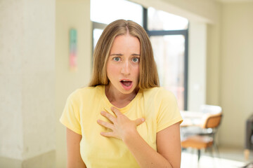 Fototapeta premium pretty blond woman feeling shocked, astonished and surprised, with hand on chest and open mouth, saying who, me?