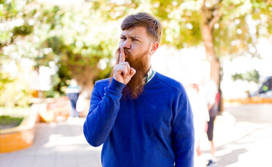 red hair bearded man looking serious and cross with finger pressed to lips demanding silence or...