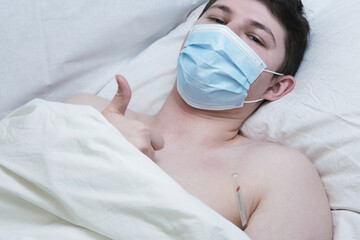 A young man lying in bed with a medical mask on his face and a thermometer under his arm, giving a...