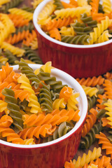 Three-colored uncooked pasta in the red ceramic bowls on the wooden background with scattered pasta on it.