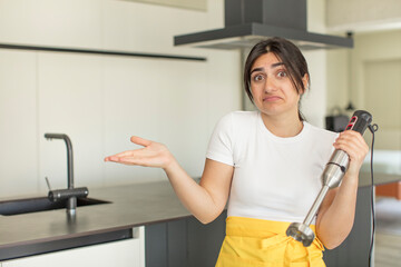 young woman shrugging, feeling confused and uncertain. chef with hand blender