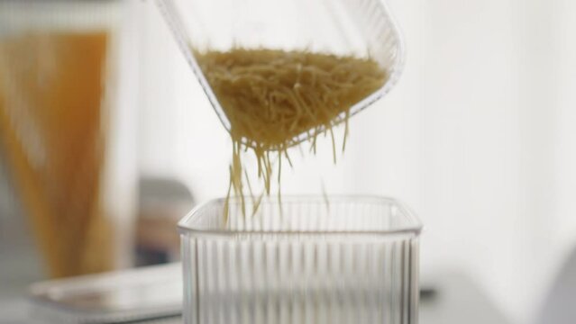 Person pours broken short spaghetti into convenient plastic container measuring portion. Preparing for cooking in cozy light kitchen. Storage of raw products