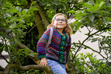 Little preschool girl with eyeglasses climbing on tree on family backyard. Lovely happy child...