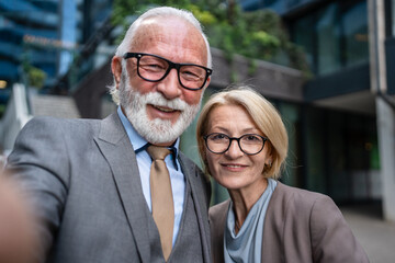 Portrait of senior man dressed in a suit and a mature blonde woman as business partners husband and wife friends or colleagues stand in front of modern building in day