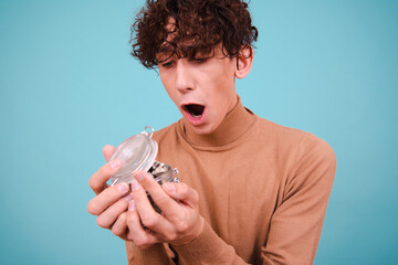 Funny curly student saves money for studying. Attractive guy posing on a blue background. - obrazy, fototapety, plakaty