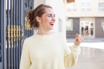 Fototapeta premium pretty young woman smiling cheerfully, feeling happy and pointing to the side and upwards, showing object in copy space