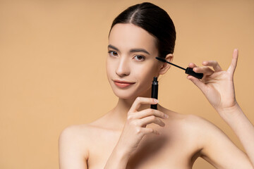 Young asian woman with naked shoulders holding mascara and applicator isolated on beige