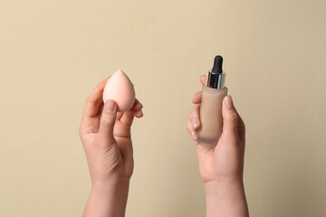 Woman holding bottle with skin foundation and makeup sponge on beige background, closeup