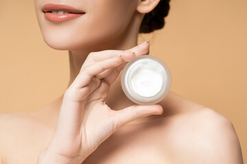 Cropped view of smiling young woman with naked shoulders holding cosmetic cream isolated on beige